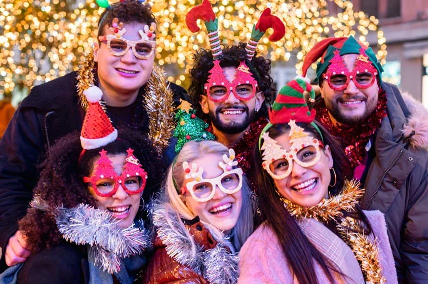 Friends smiling and posing together at a festive holiday-themed party while wearing Christmas glasses and decorations.